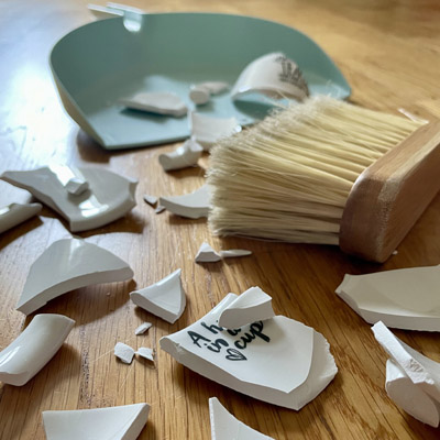 Symbol Eifersucht: Eine zerbrochene Tasse auf dem Boden. Daneben ein Handfeger und eine Kehrschaufel.